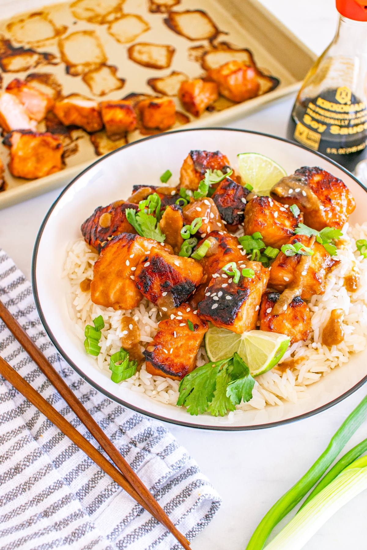 A bowl of rice topped with glazed salmon, green onions, cilantro, lime wedges, and sesame seeds.