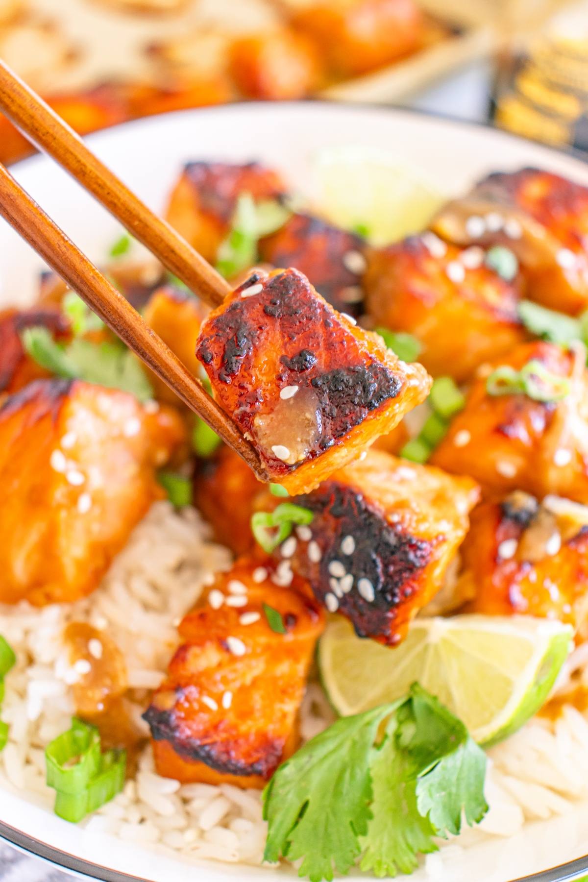 Chopsticks hold a glazed, grilled salmon bite over a bowl of rice, herbs, and lime slices.