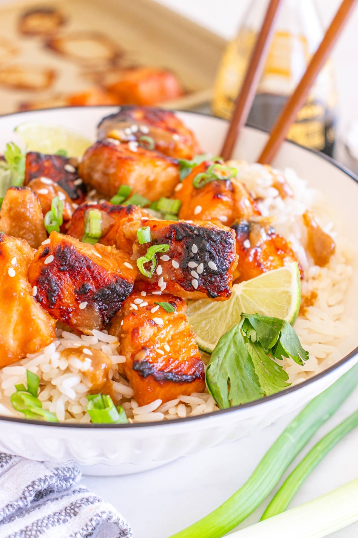 Grilled glazed salmon over rice, garnished with green onions, sesame seeds, lime wedges, and cilantro.