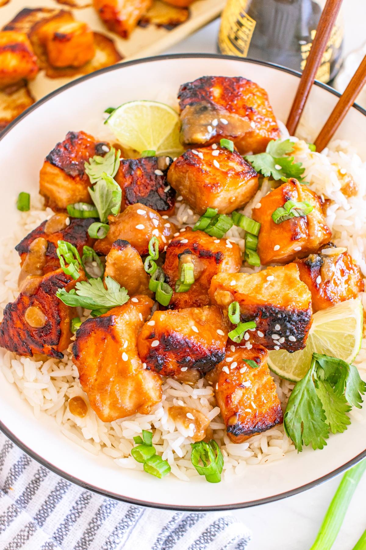 A bowl of white rice topped with glazed grilled salmon, green onions, cilantro, lime slices, and sesame seeds.