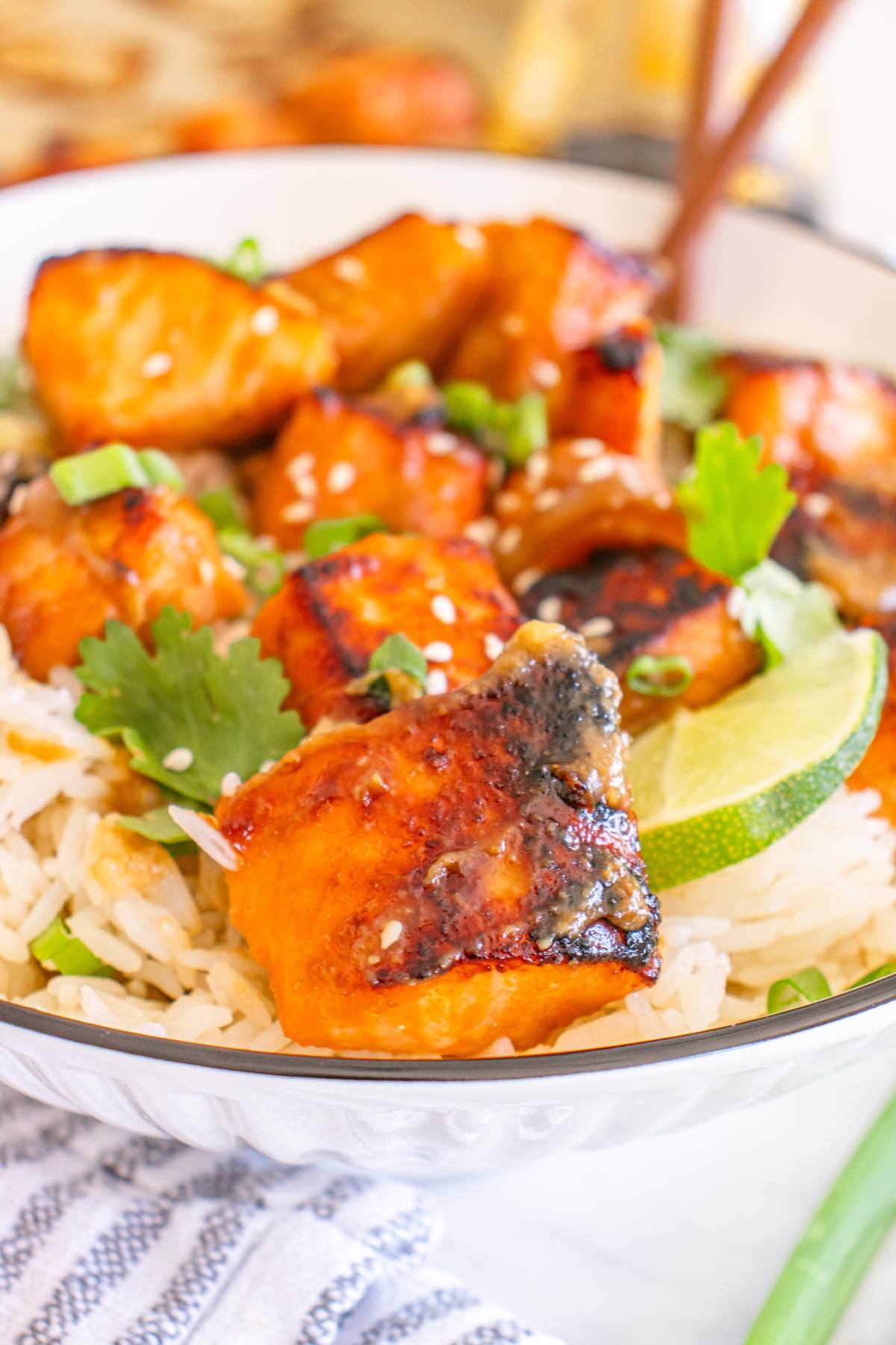 Bowl of grilled salmon over rice, topped with sesame seeds, green onions, cilantro, and a lime wedge.