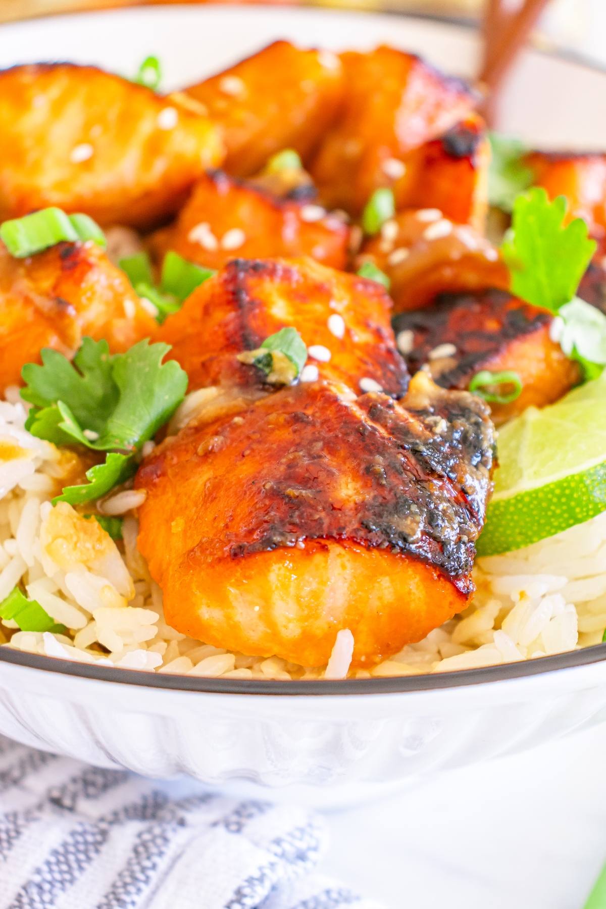 Grilled salmon pieces on rice, garnished with sesame seeds, cilantro, green onions, and lime wedges.
