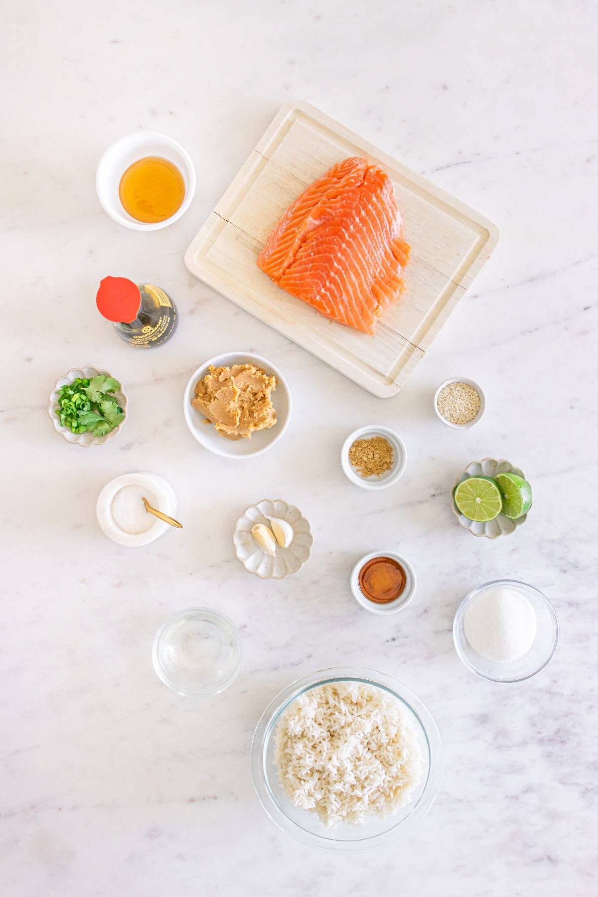 Overhead view of raw salmon, rice, and various sauces, spices, and herbs arranged on a white countertop.