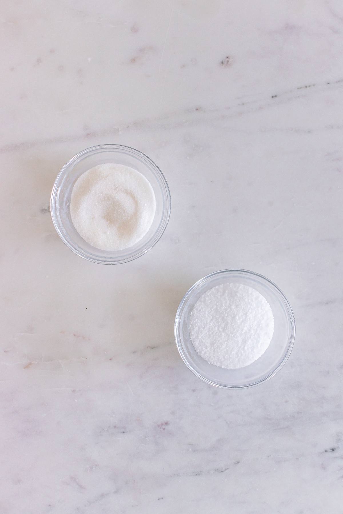 Two small glass bowls, each filled with white granules, sit on a white marble surface.