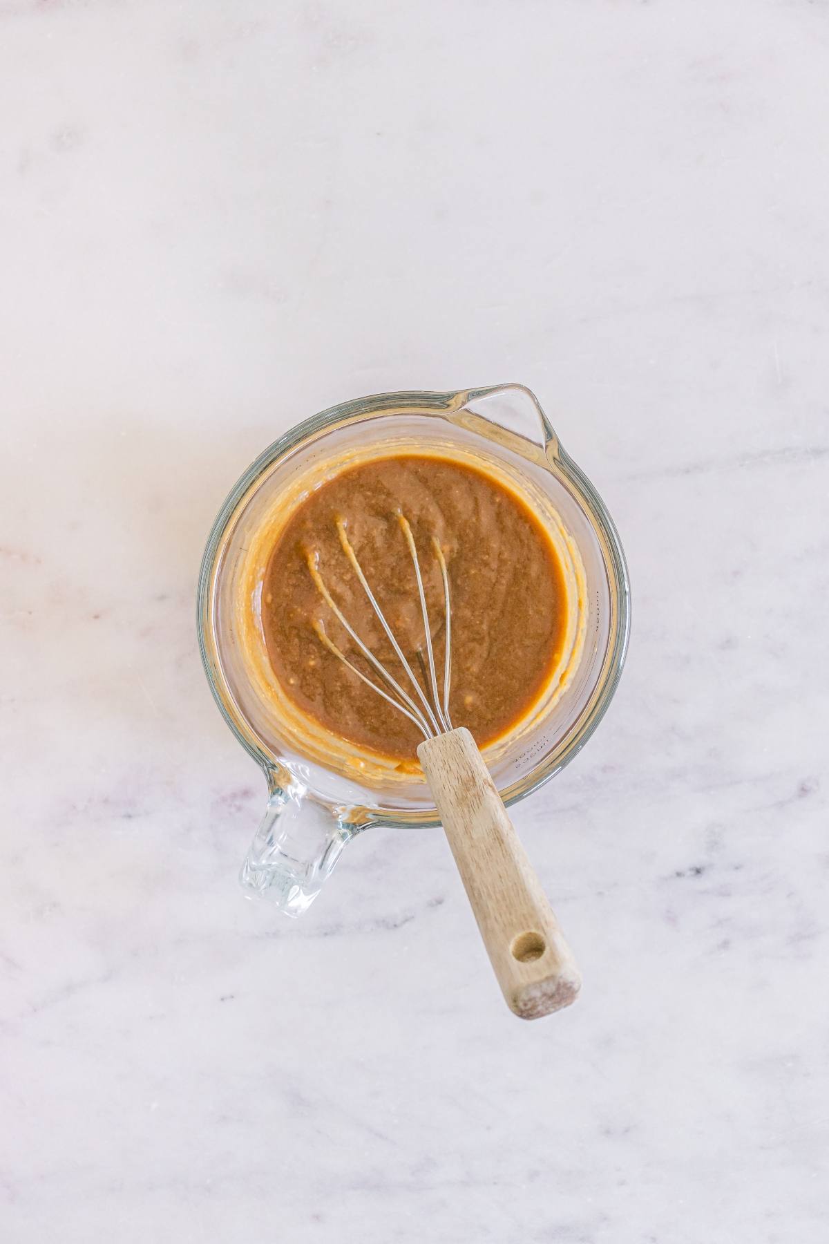 Glass mixing bowl with a whisk, containing a thick brown batter, on a light marble surface.