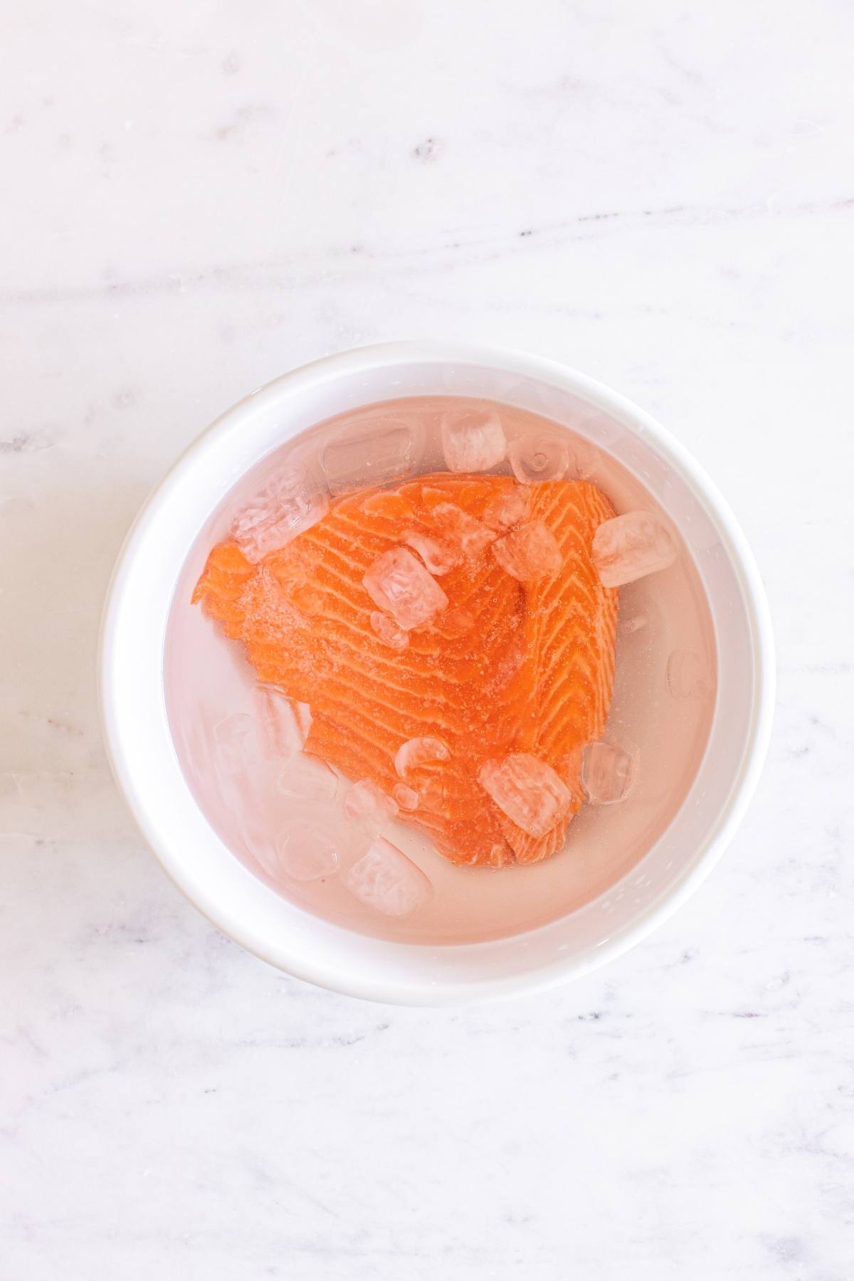 A raw salmon fillet in a white bowl filled with ice cubes and water, on a light marble surface.