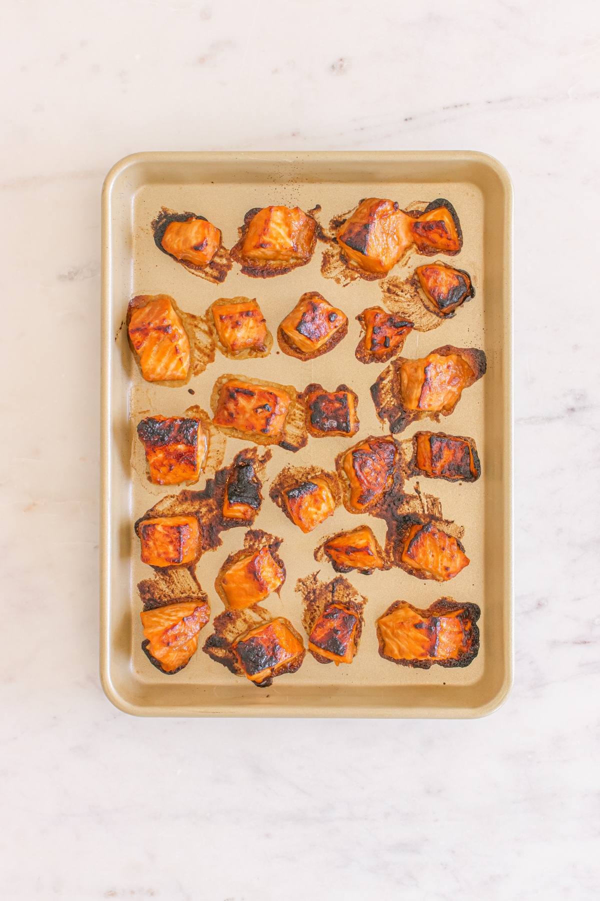 Baked salmon bites on a gold sheet pan, arranged on a light marble countertop.