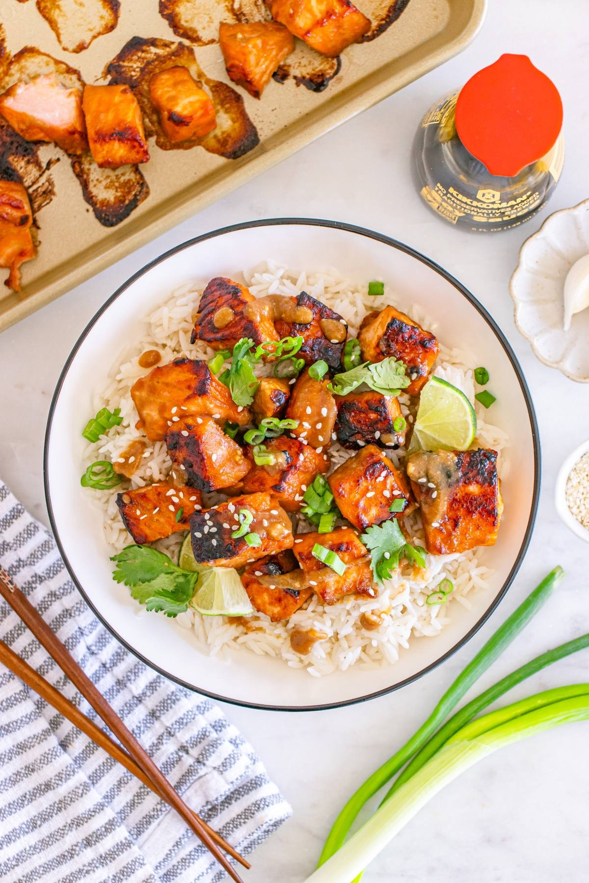 A bowl of white rice topped with glazed salmon bites, garnished with sesame seeds, green onions, and lime.