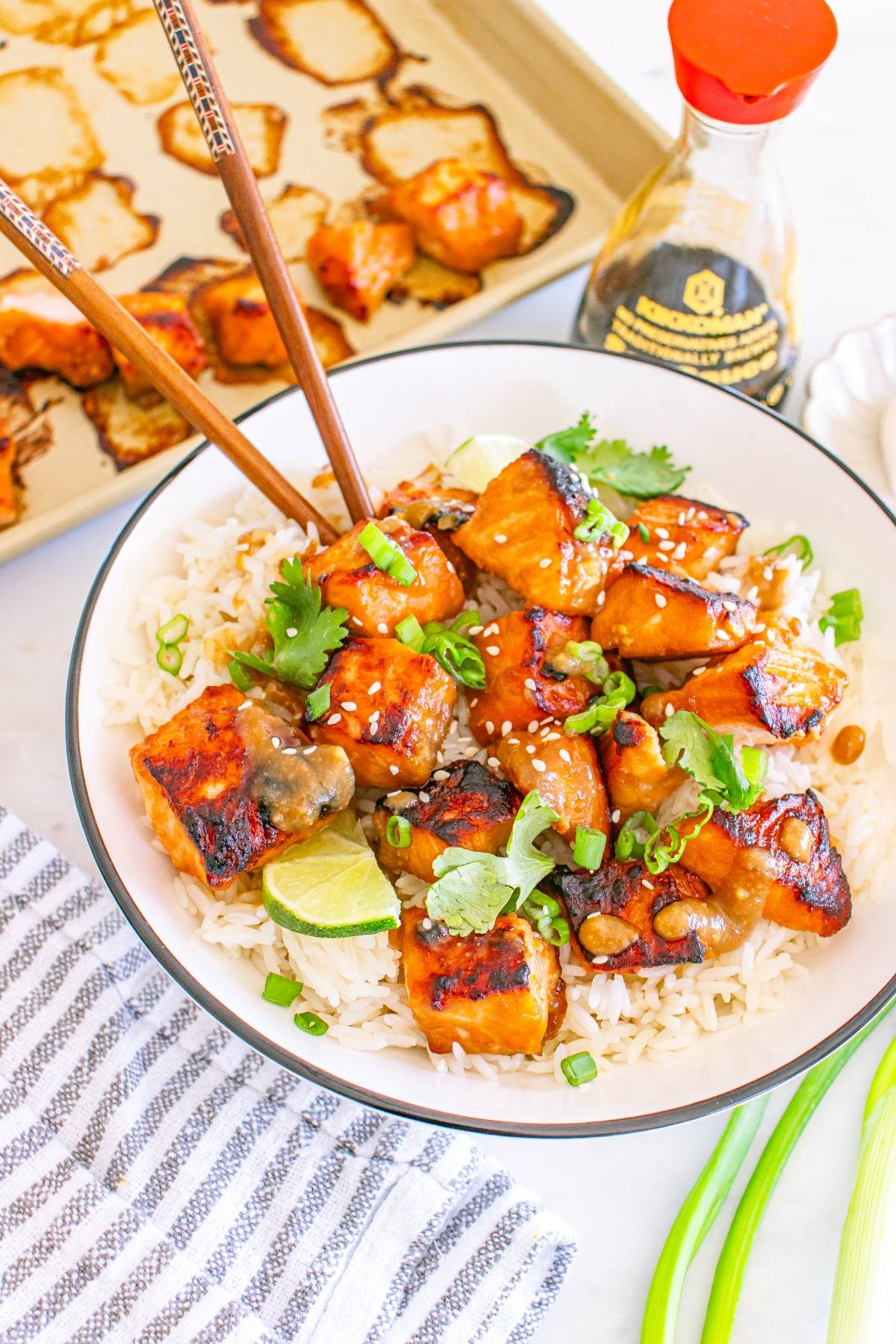 A bowl of rice topped with glazed tofu, cilantro, green onions, and sesame seeds, with chopsticks and lime wedges.