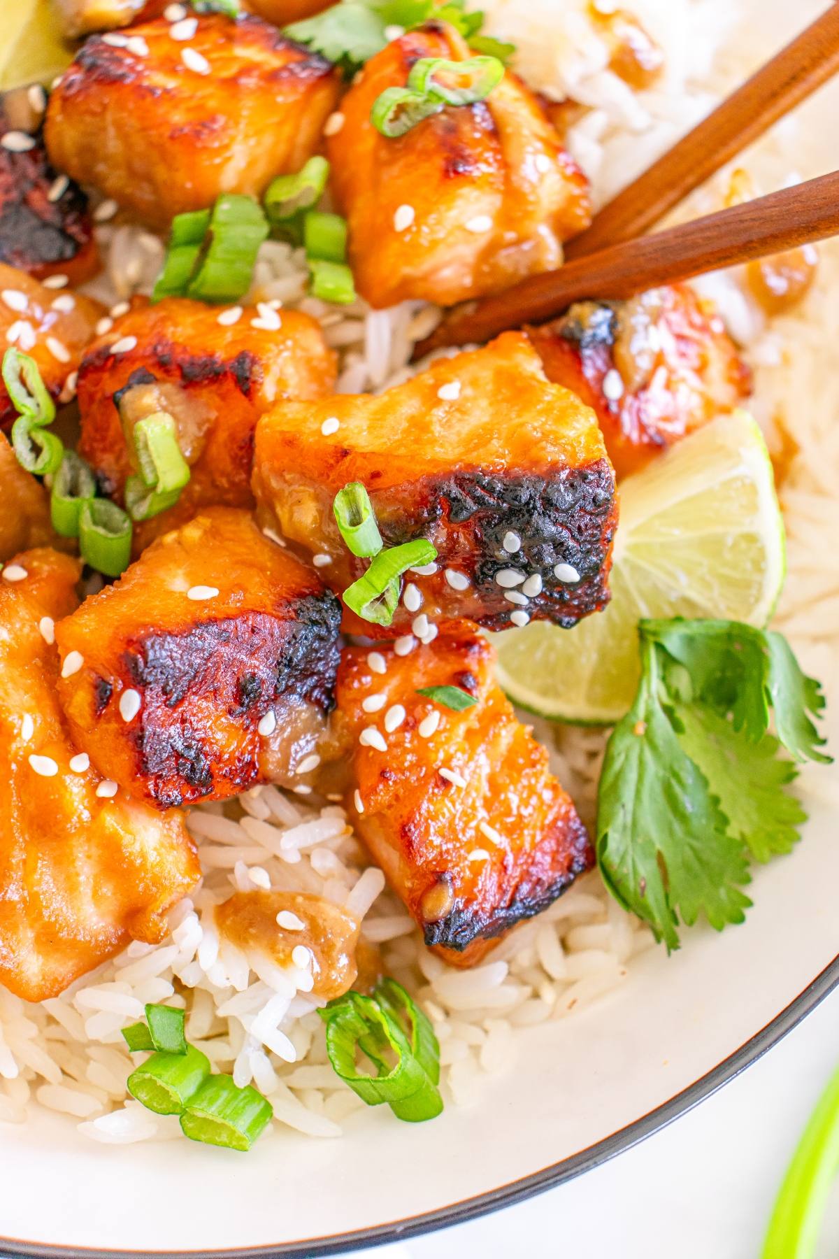 Grilled chicken on rice with green onions, sesame seeds, lime wedges, and cilantro, with chopsticks.