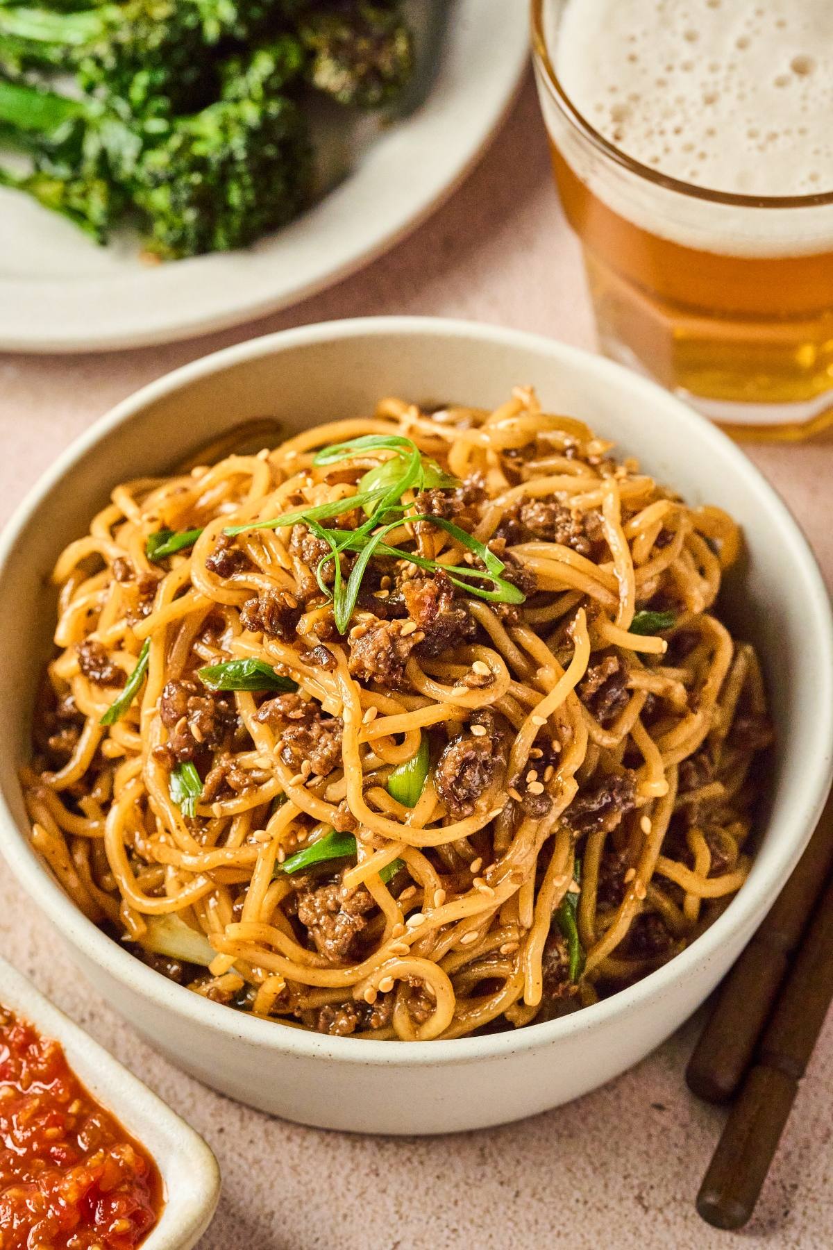 A bowl of saucy noodles with ground meat, garnished with green onions, next to a drink and vegetables.