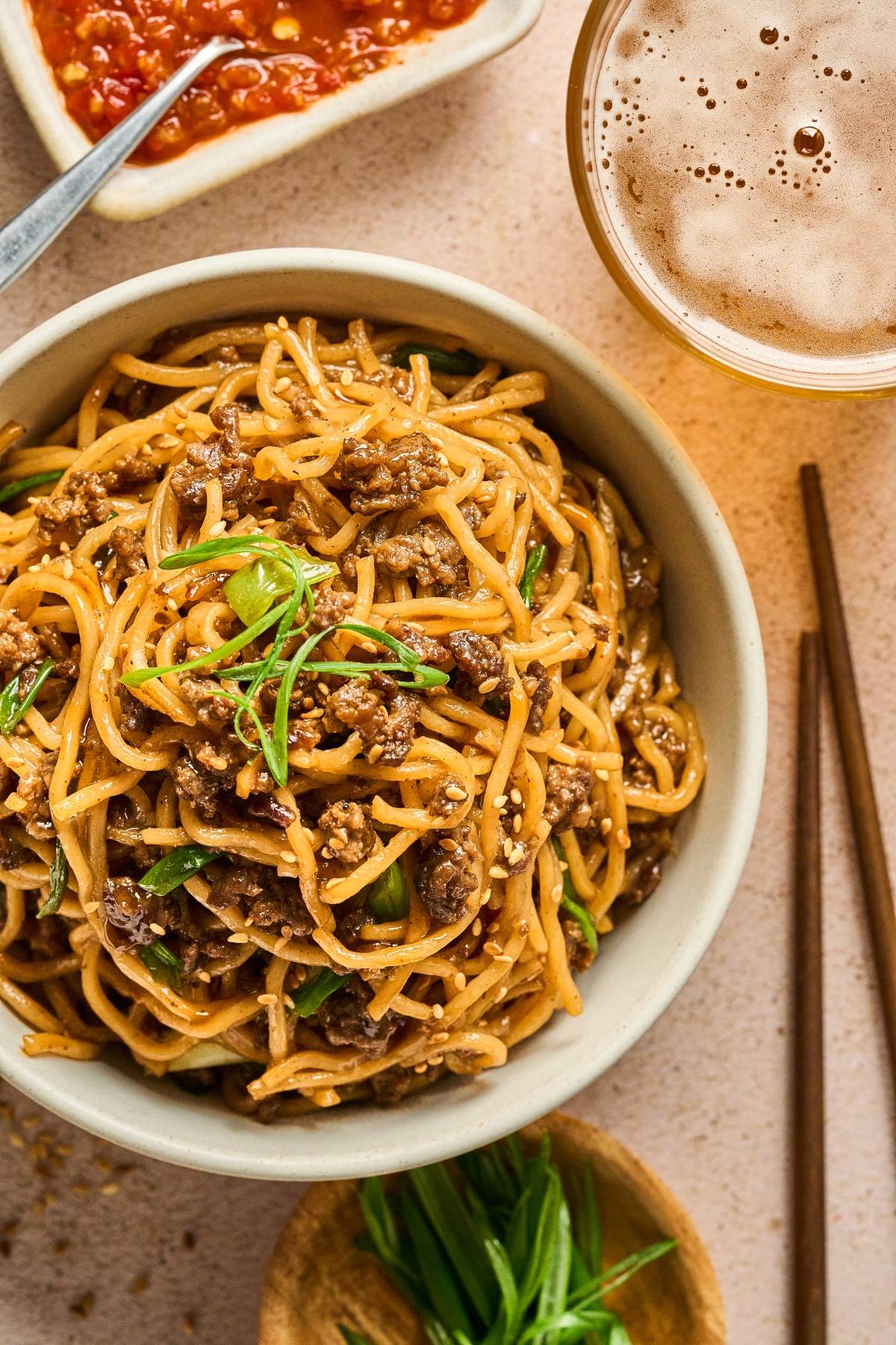 Bowl of stir-fried noodles with ground meat and scallions, with chopsticks, sauce, and a drink nearby.