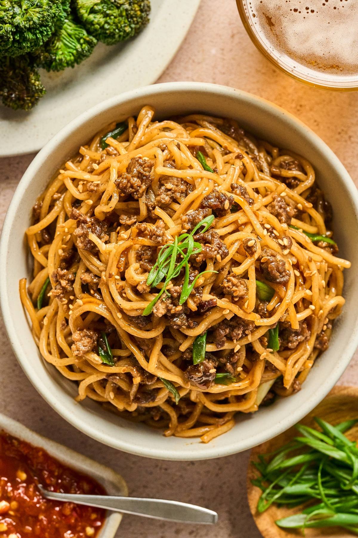 A bowl of noodles with ground meat and green onions, surrounded by broccoli, sauce, and a drink.