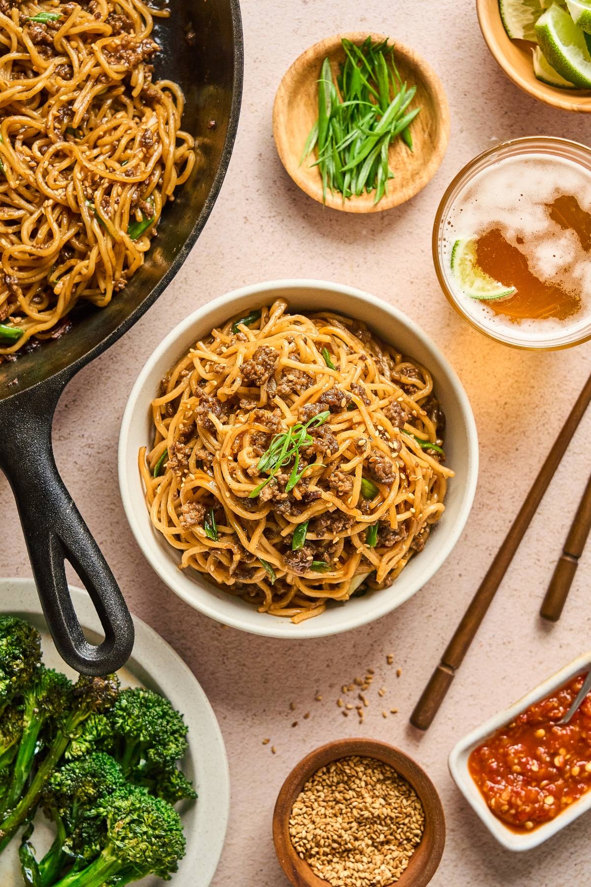 Bowl of noodles with minced meat, scallions, and chopsticks, surrounded by broccoli, beer, and sauces.