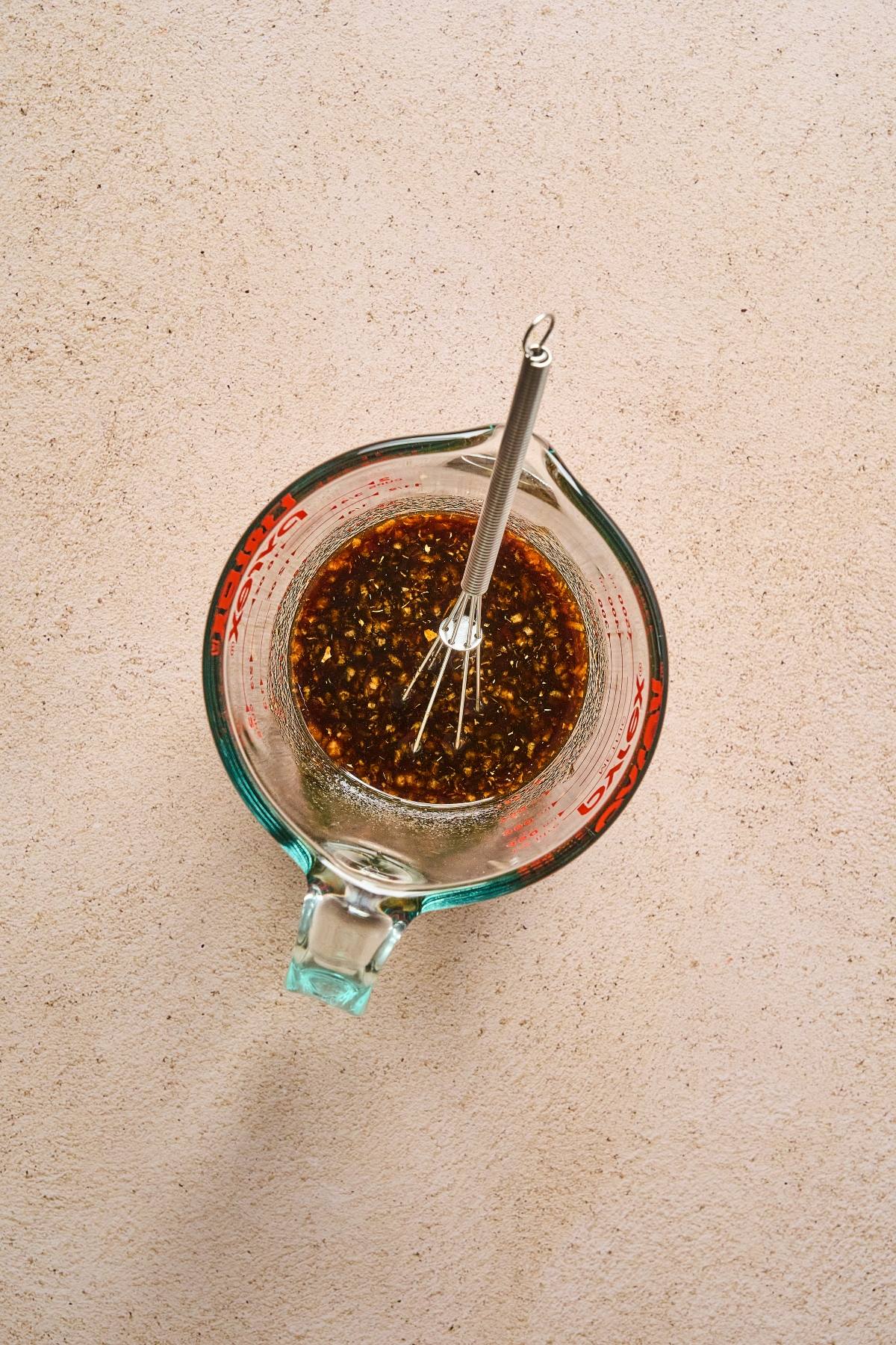 A glass measuring cup with a metal whisk and a dark, speckled sauce inside, on a light countertop.