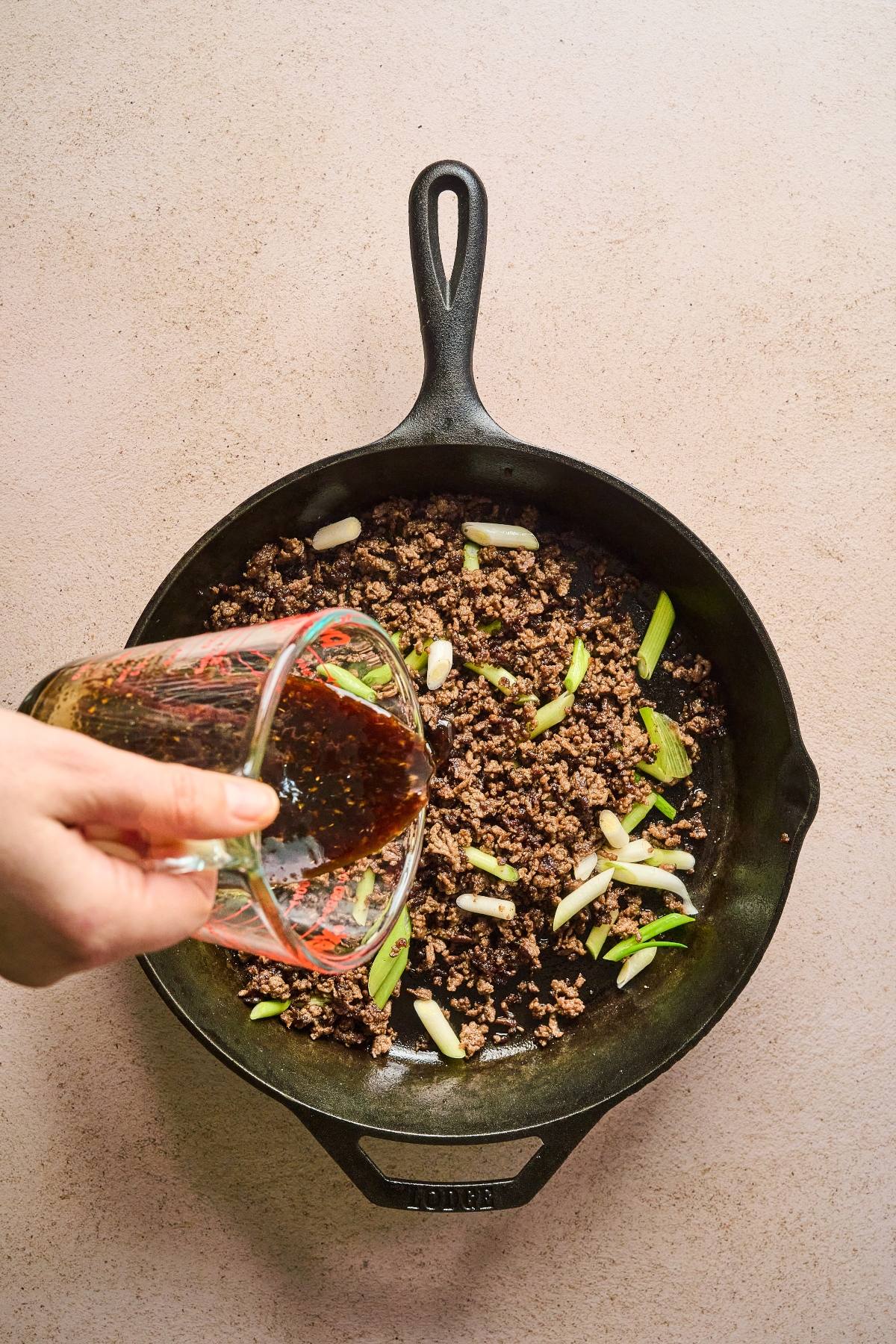 A hand pours sauce into a skillet with cooked ground meat and green onions on a light countertop.