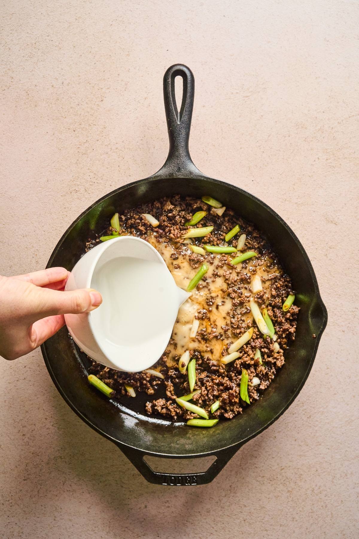 A hand pours milk into a skillet with cooked ground beef and sliced green onions.