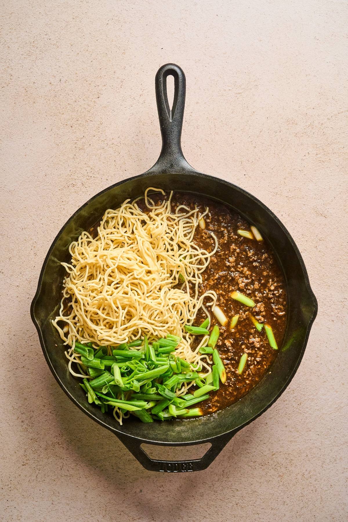 A cast iron skillet with noodles, green onions, and minced meat in sauce on a beige surface.