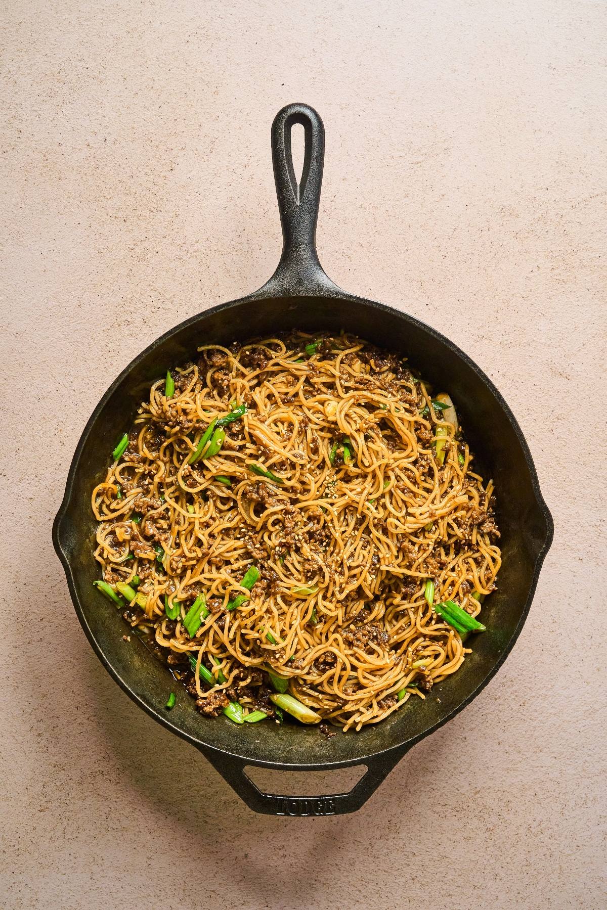 A cast iron skillet filled with stir-fried noodles, ground meat, and green onions on a light surface.