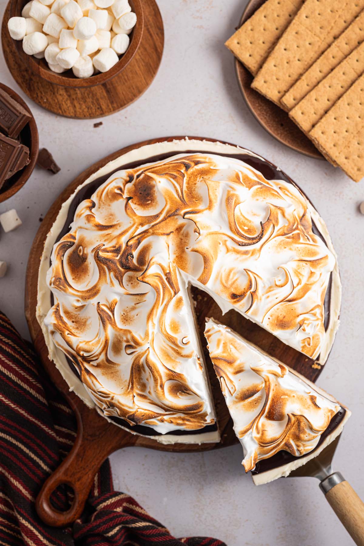 A toasted s'mores pie with a slice being served, surrounded by marshmallows, chocolate, and graham crackers—a tempting scene reminiscent of a No Bake S'mores Cheesecake.