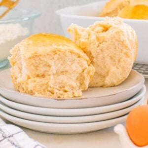 Two fluffy Parker House Rolls rest on stacked plates with eggs, flour, and a baking dish in the background.