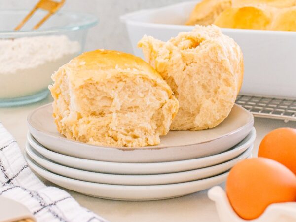 Two fluffy Parker House Rolls rest on stacked plates with eggs, flour, and a baking dish in the background.