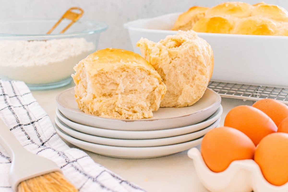 Two homemade Parker House Rolls on stacked plates, with eggs, flour, and a baking dish in the background.
