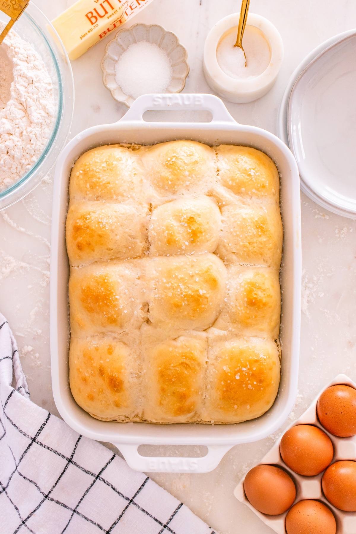 A baking dish of golden brown dinner rolls on a countertop with eggs, butter, flour, and a towel nearby.