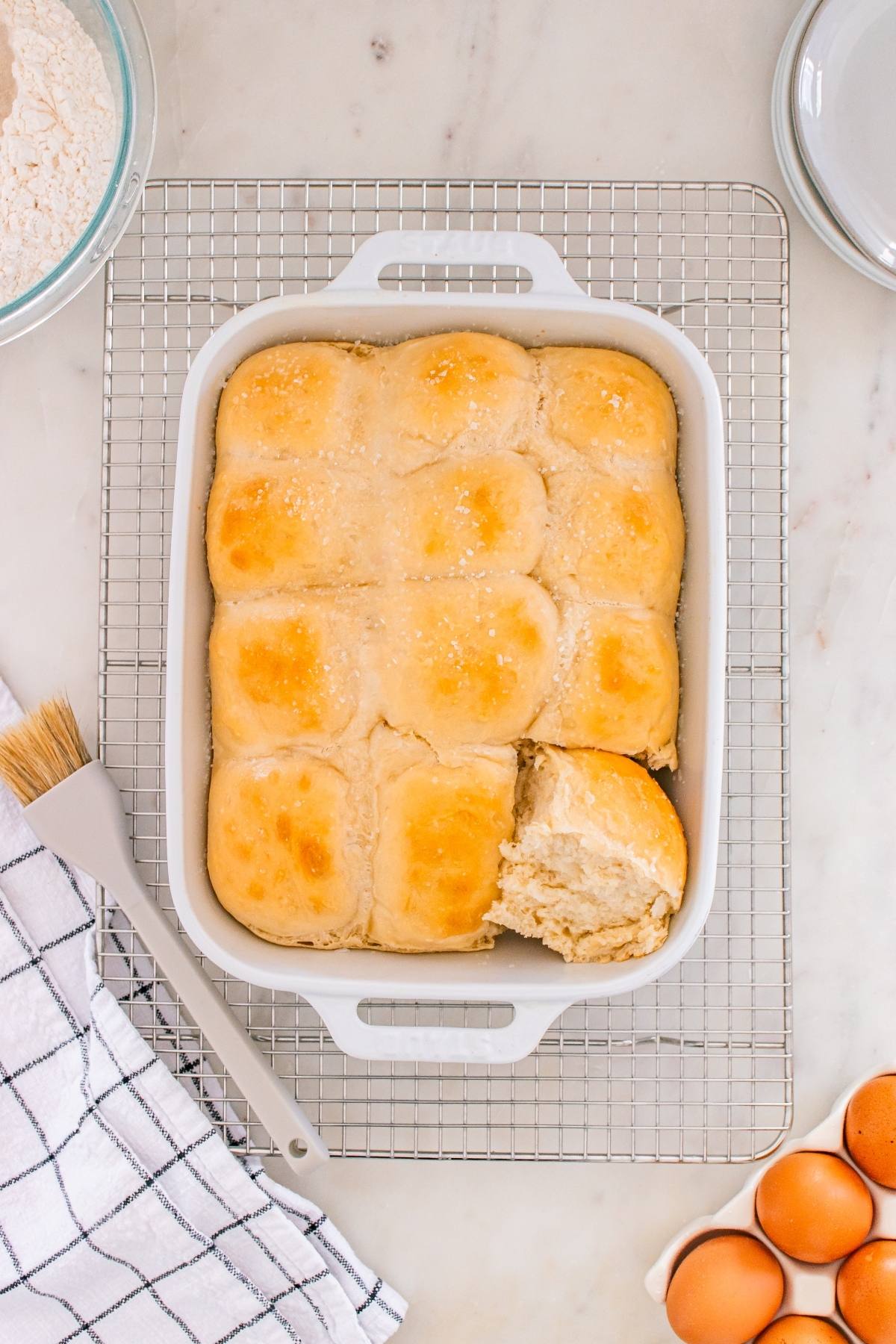 A pan of golden brown dinner rolls, one partially removed, on a cooling rack with baking items nearby.