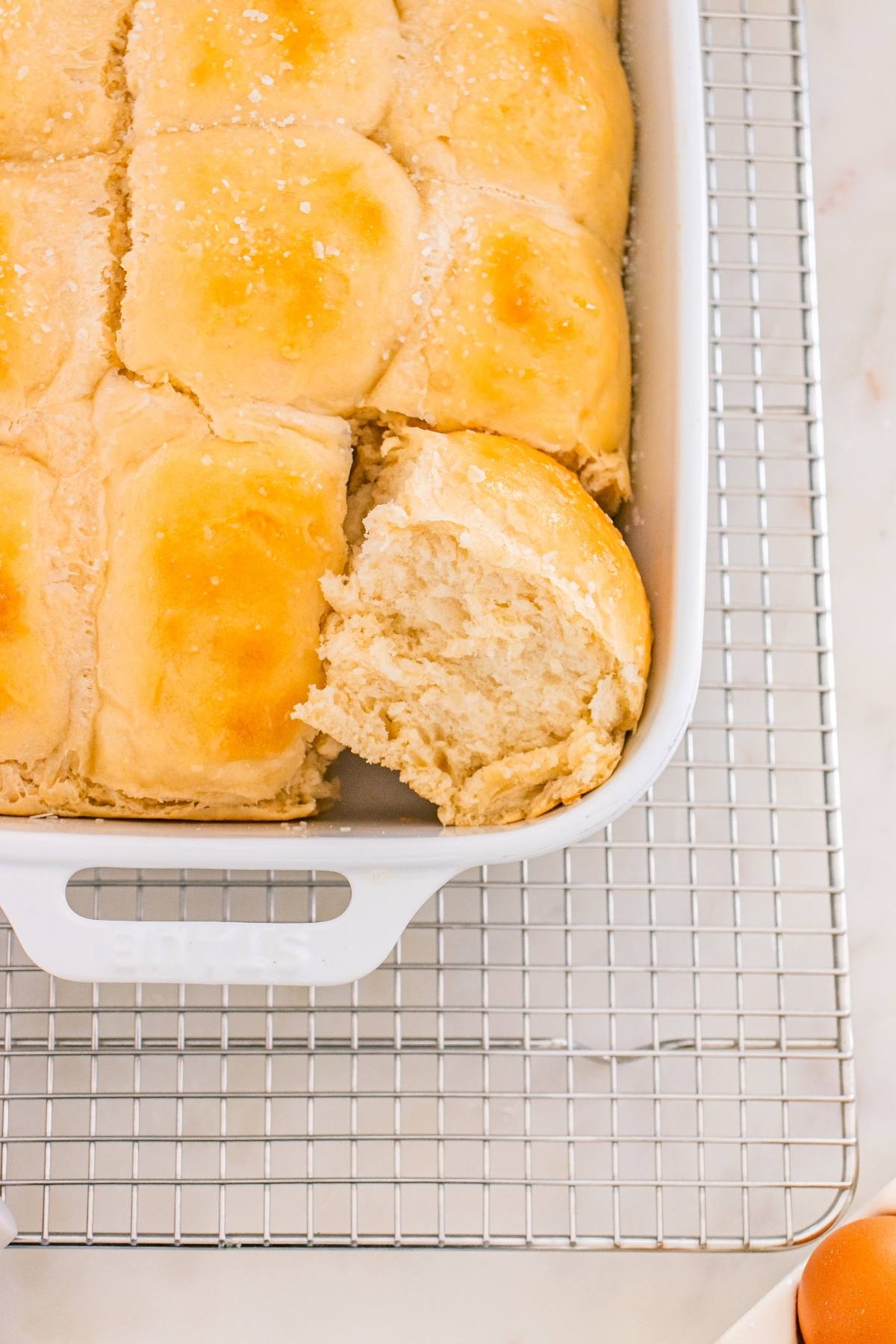 A baking dish of golden brown dinner rolls, with one roll pulled out, on a cooling rack.