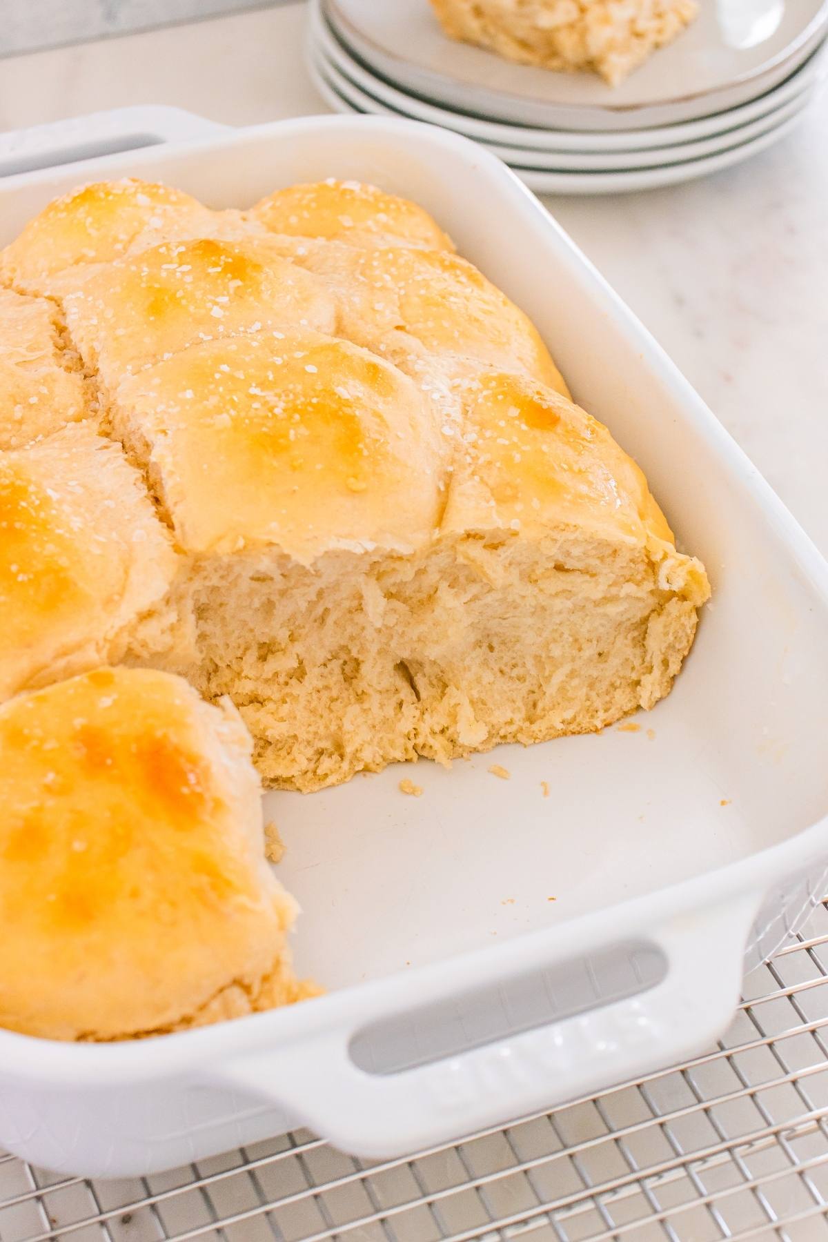 Golden brown dinner rolls in a white baking dish, with one roll missing and visible flaky texture inside.