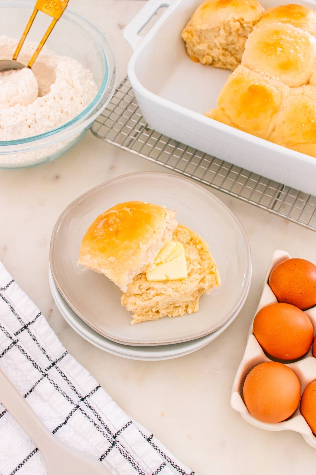 A buttered biscuit sits on a plate next to eggs, flour, and a dish of freshly baked biscuits.