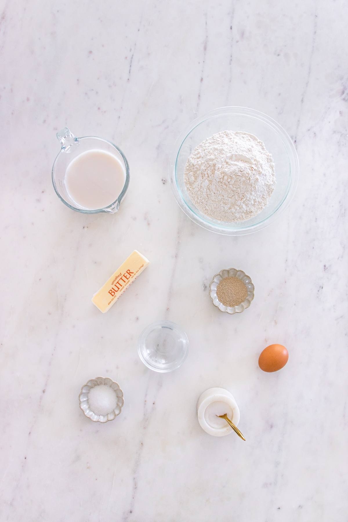 Baking ingredients on marble: milk, flour, butter, yeast, egg, salt, and sugar in small containers.