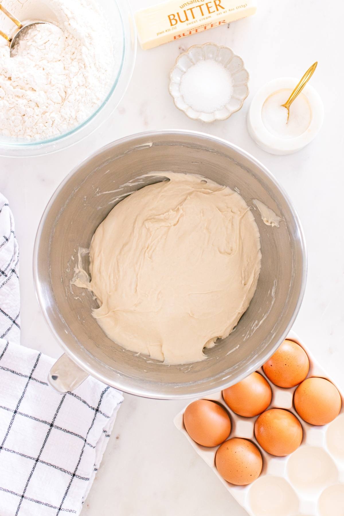 Metal mixing bowl with dough, eggs in a tray, butter, flour, salt, sugar, and a kitchen towel on a counter.