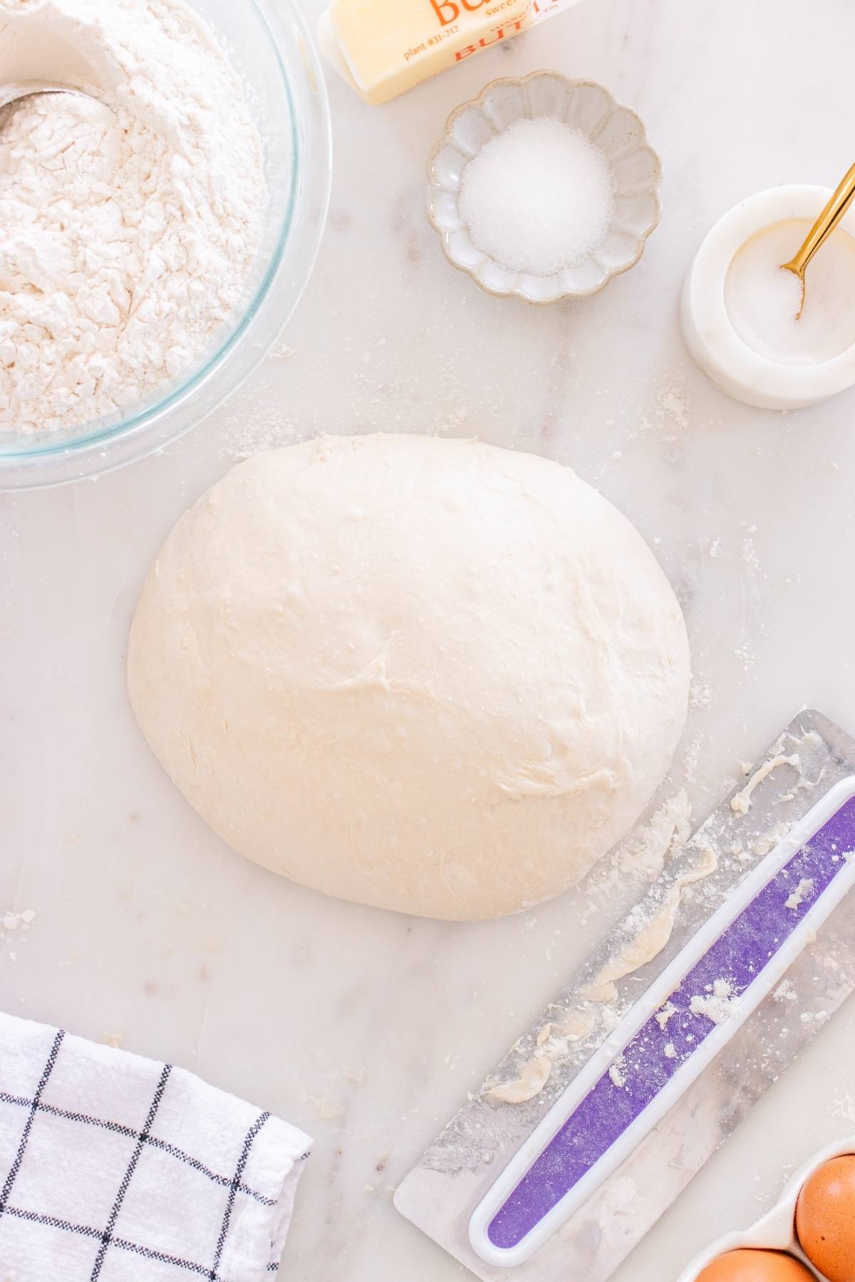 A ball of bread dough on a floured surface with flour, sugar, a scraper, and eggs nearby.