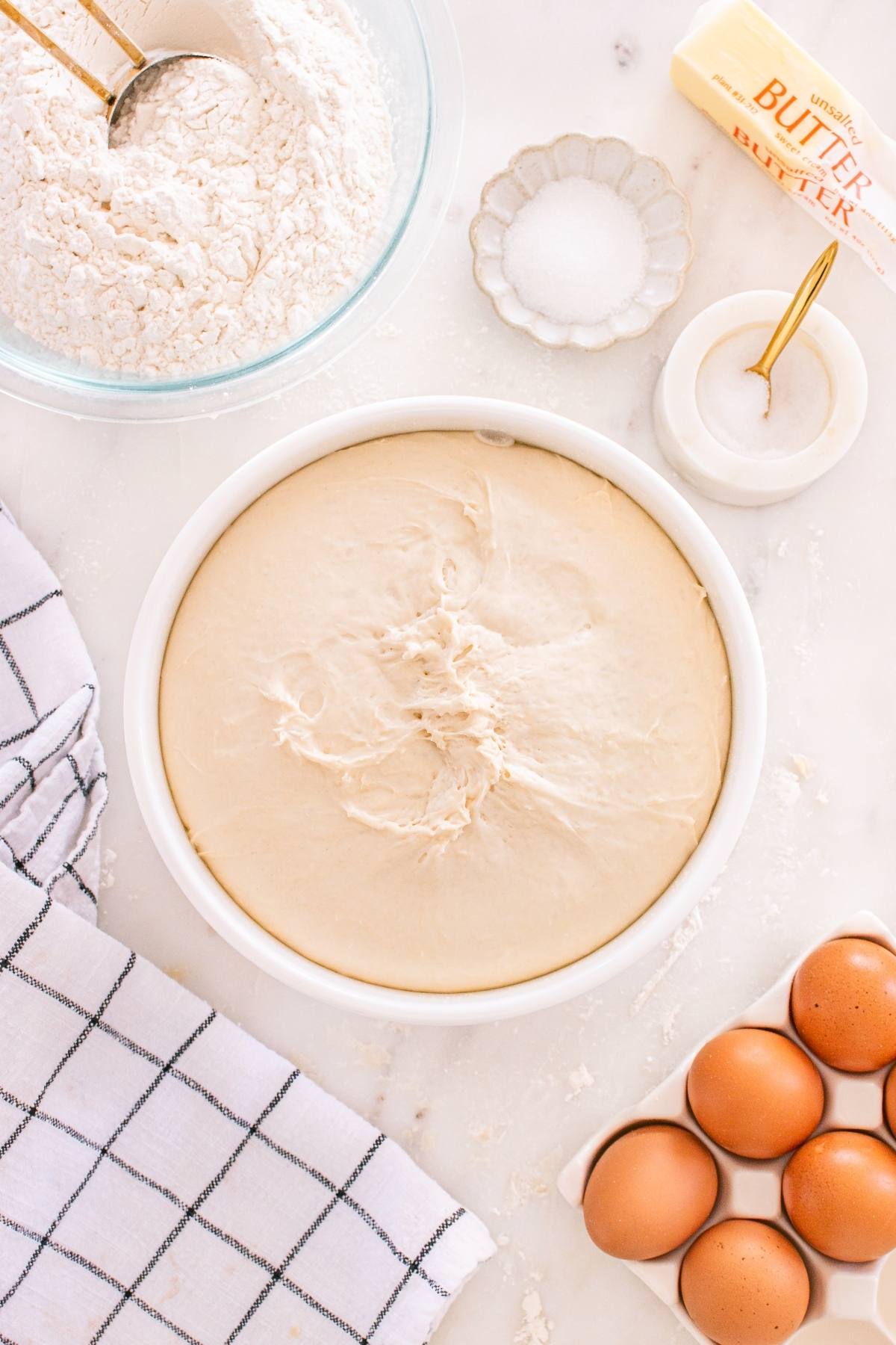 A bowl of risen dough surrounded by flour, eggs, butter, salt, and a striped kitchen towel on a white surface.