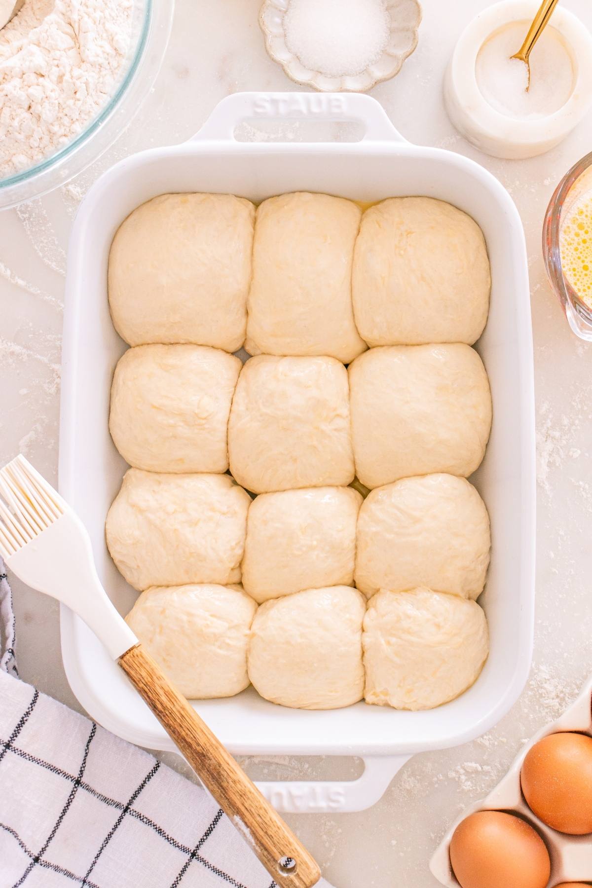 A white baking dish with twelve unbaked dough rolls, surrounded by baking ingredients and utensils.