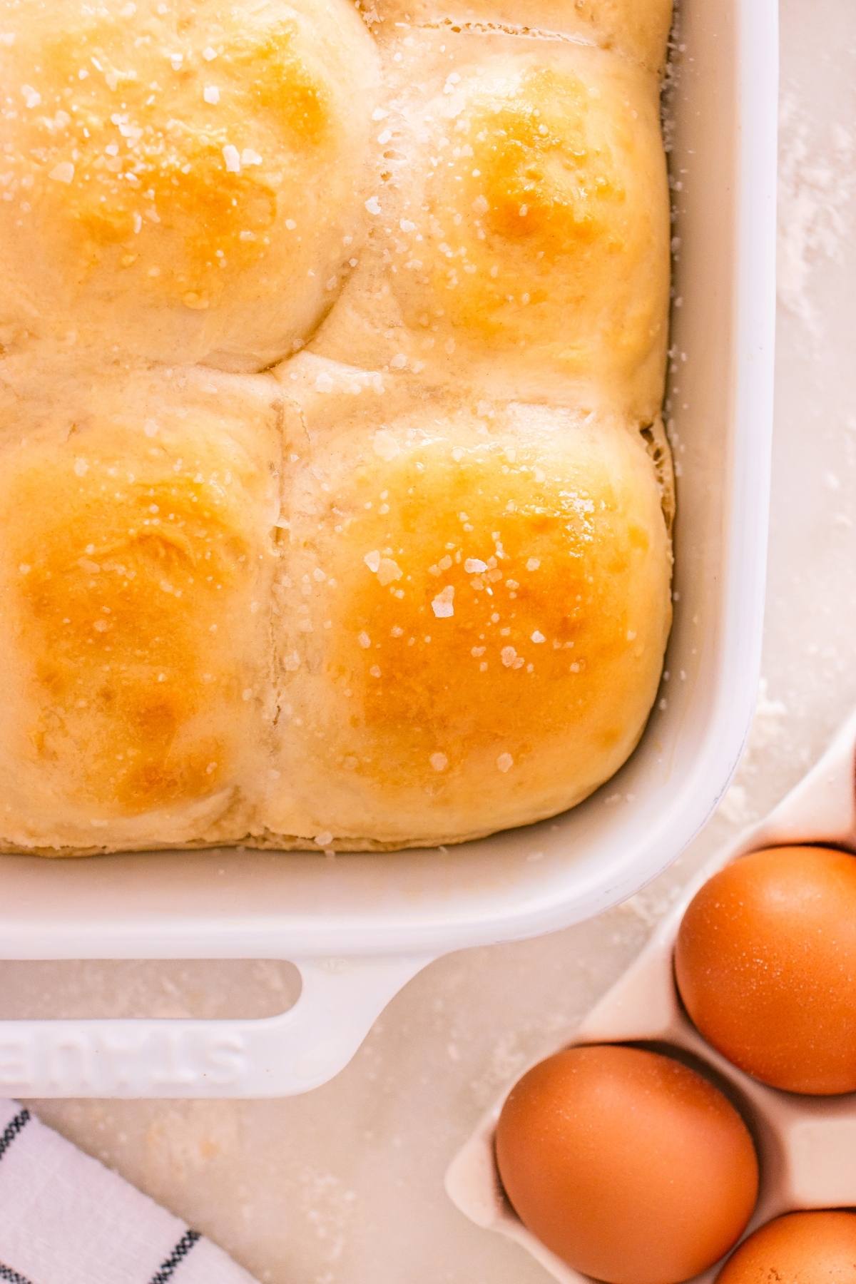 Golden dinner rolls topped with coarse salt in a baking dish, next to brown eggs on a light surface.