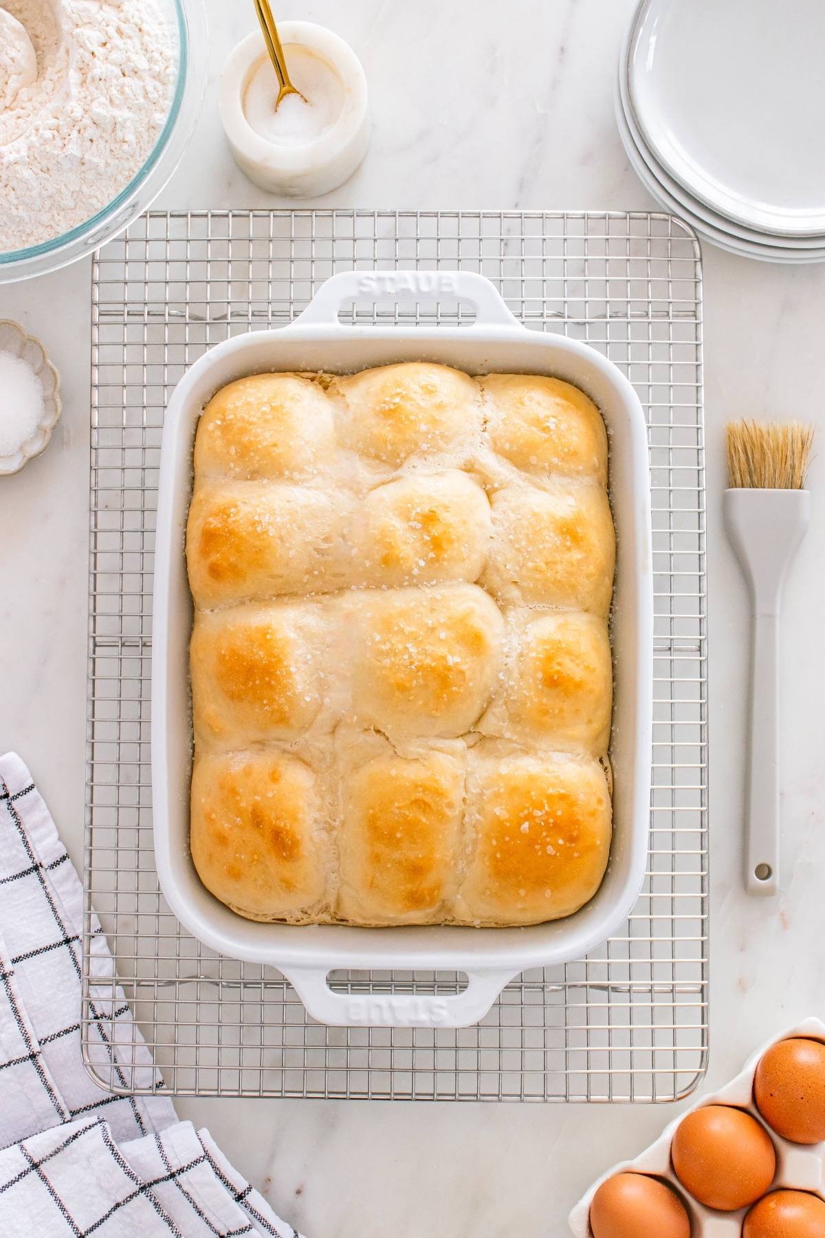 Freshly baked golden dinner rolls in a white baking dish on a cooling rack, surrounded by baking tools.