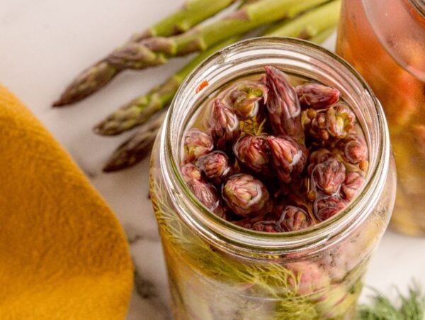 Open jar of Pickled Asparagus with fresh dill, beside whole asparagus spears and a yellow cloth for a vibrant, tangy display.