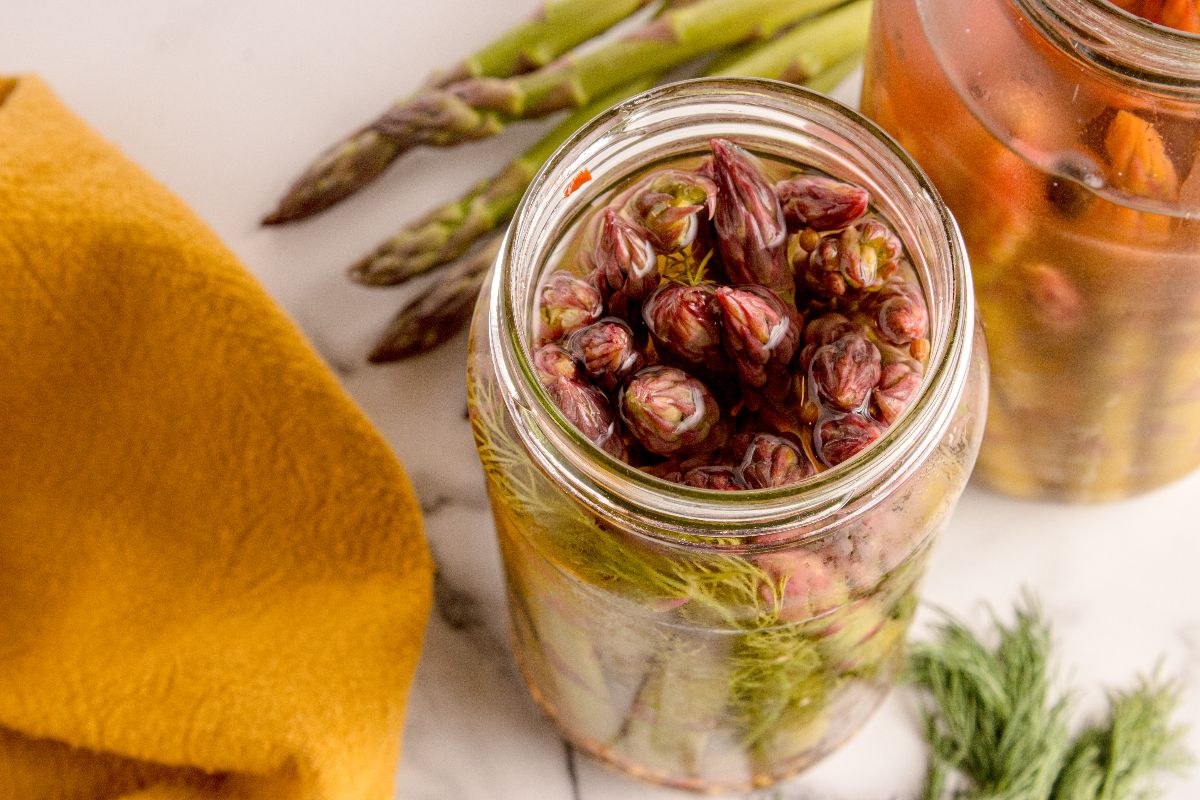 A jar of Pickled Asparagus spears sits on a white surface, with fresh asparagus and aromatic dill arranged beside it.