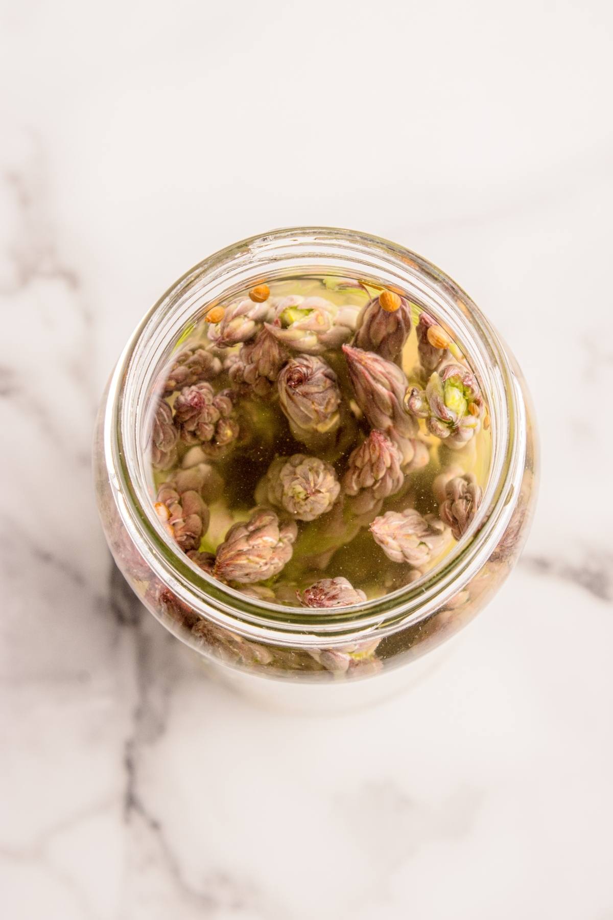 Glass jar filled with pickled asparagus tips and spices on a white marble surface.