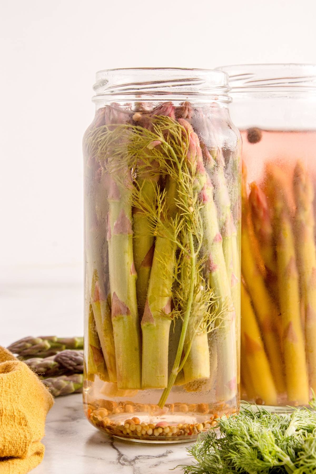 Glass jar filled with pickled asparagus spears, dill, and spices in brine on a light surface.