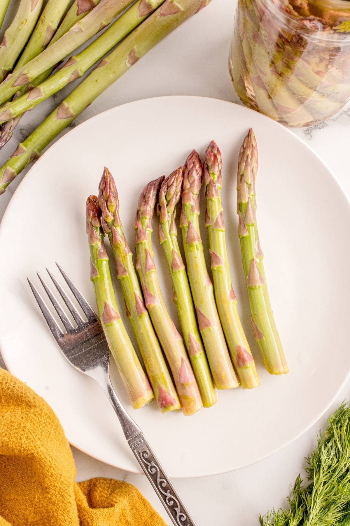 Six spears of asparagus on a white plate with a fork, next to a yellow cloth and fresh dill.