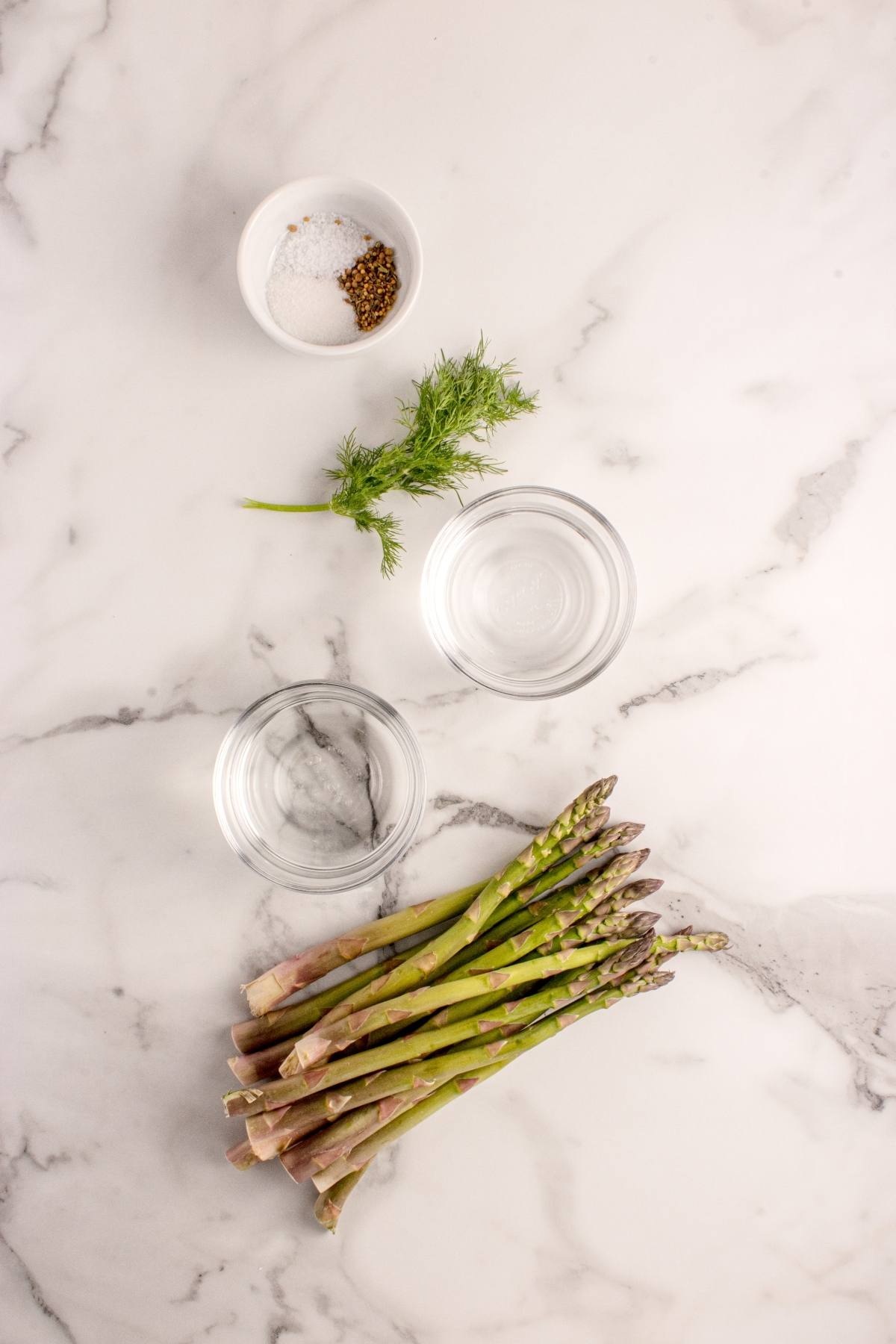Asparagus, two bowls of water, a bowl of salt and pepper, and fresh dill on a marble surface.