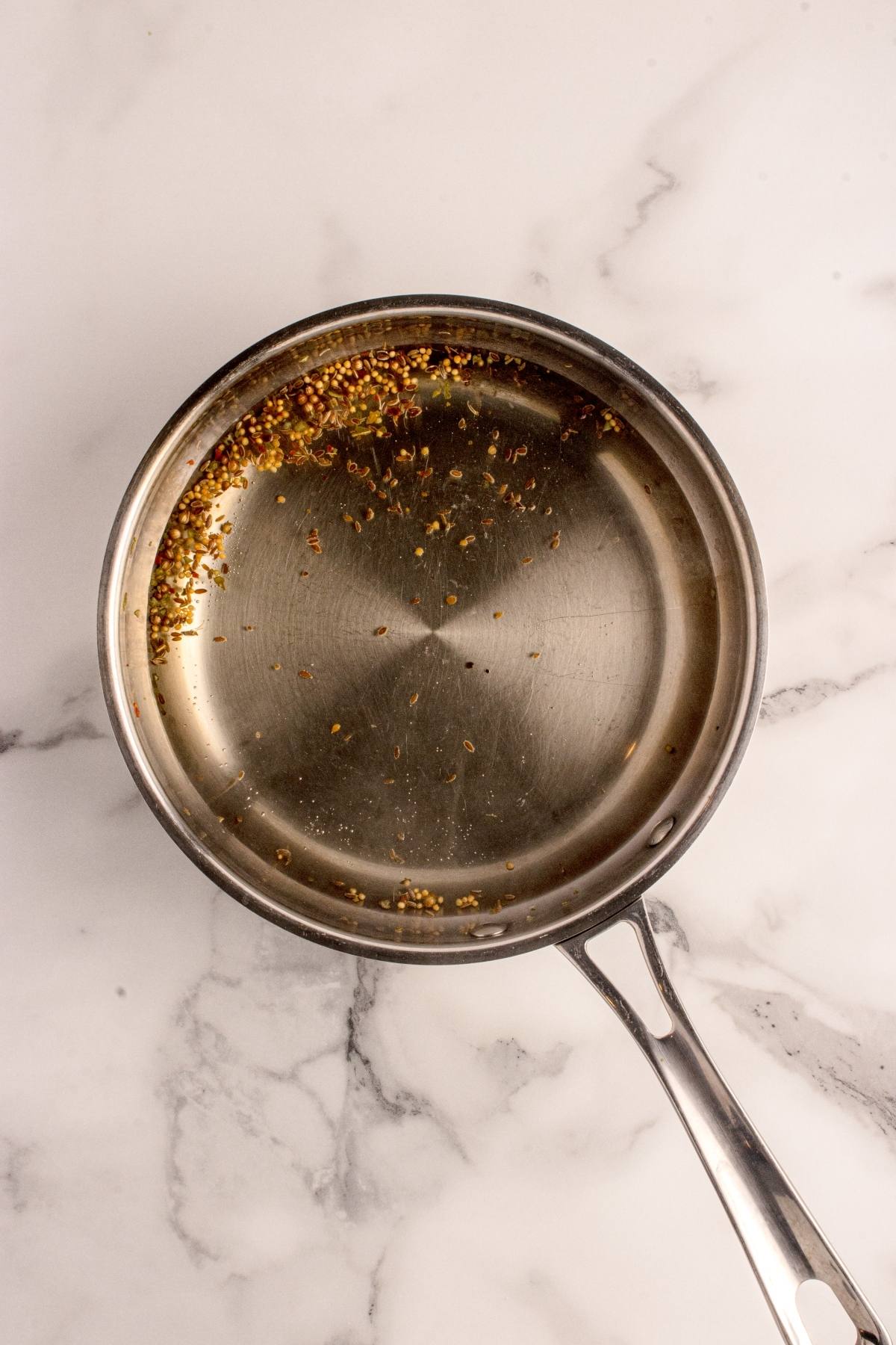 A stainless steel pan with scattered spice seeds sits on a white marble surface.