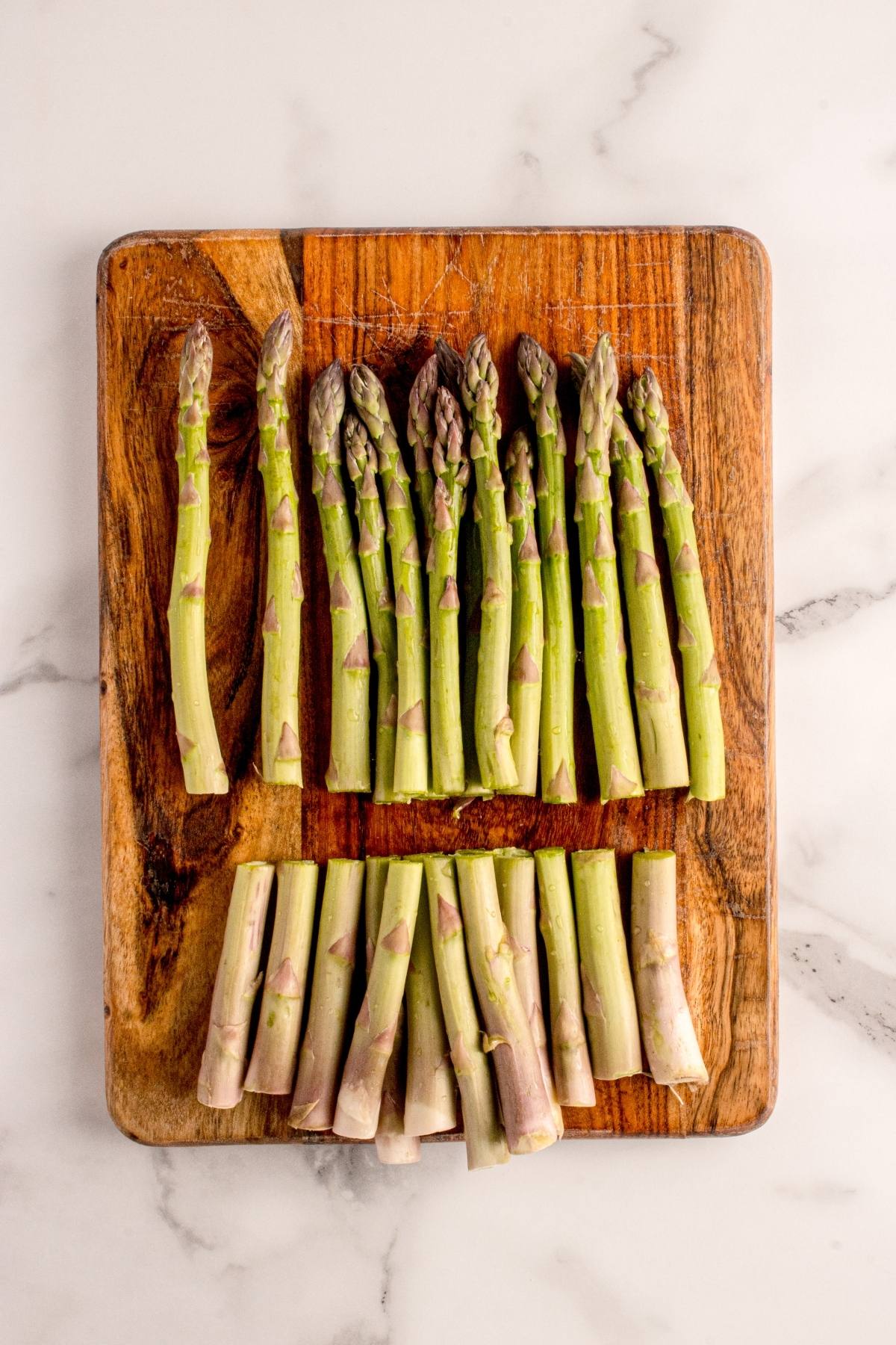 A wooden board with fresh asparagus spears, tips on top and cut stalks below, on a white marble surface.