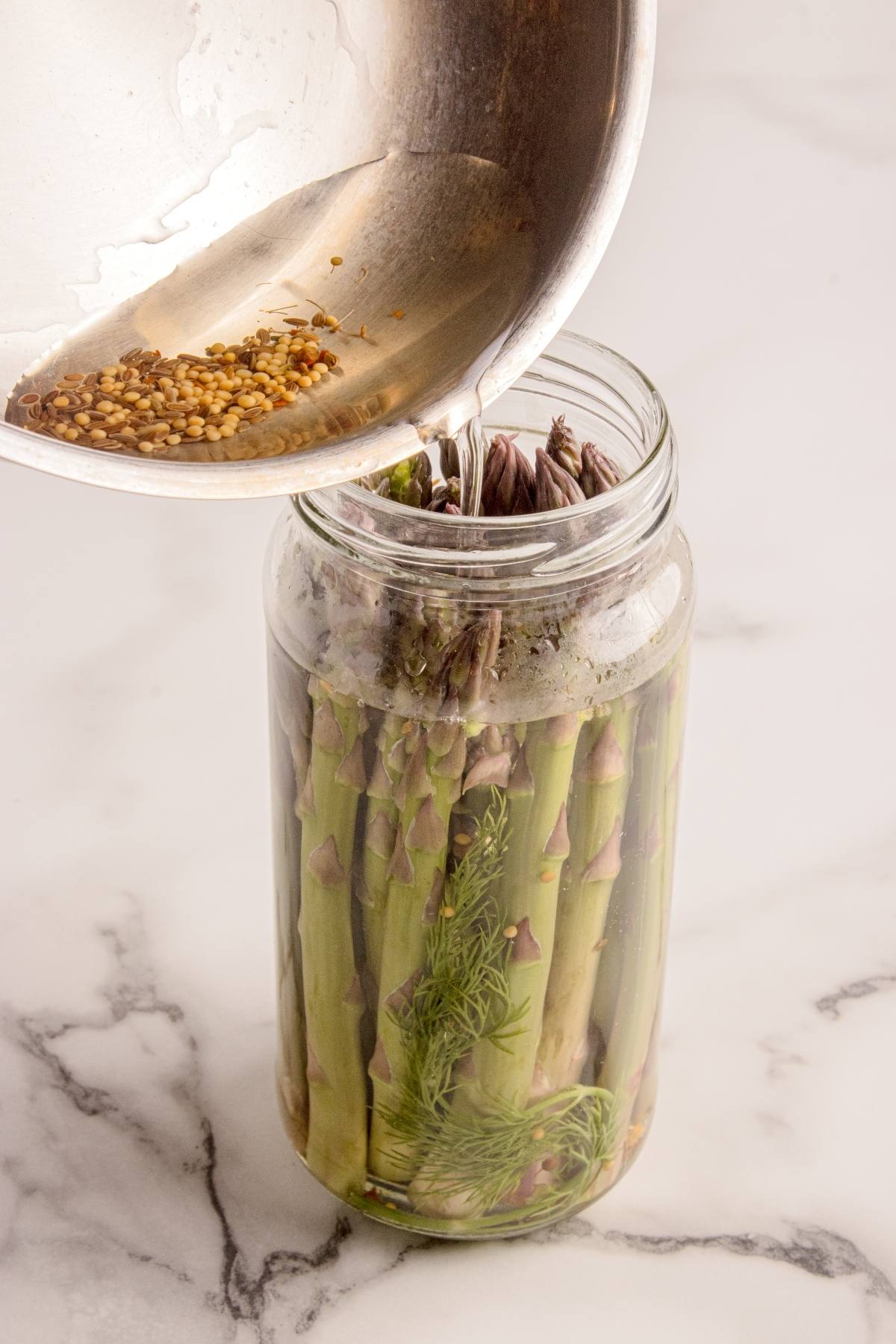 A jar of asparagus spears with dill is being covered with pickling brine from a metal bowl.