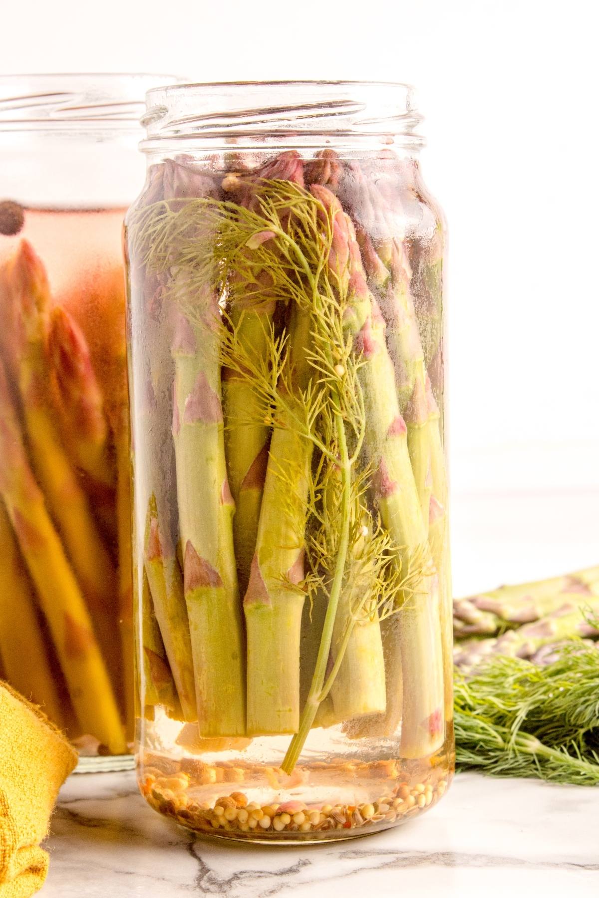 Glass jar filled with pickled asparagus, dill, and spices on a marble surface.