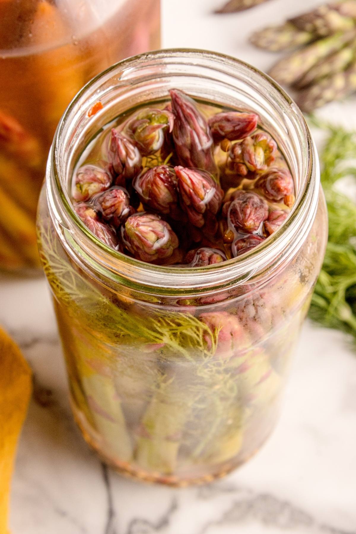 A glass jar filled with pickled asparagus spears and dill on a marble surface.